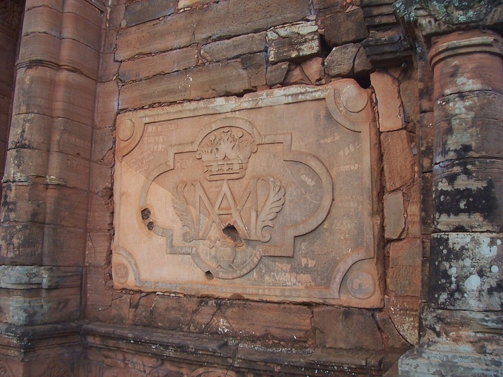 Placa de pedra desgastada com relevo de coroa e letras MA aladas, embutida em parede de tijolos avermelhados e colunas laterais, com musgo e pichações
