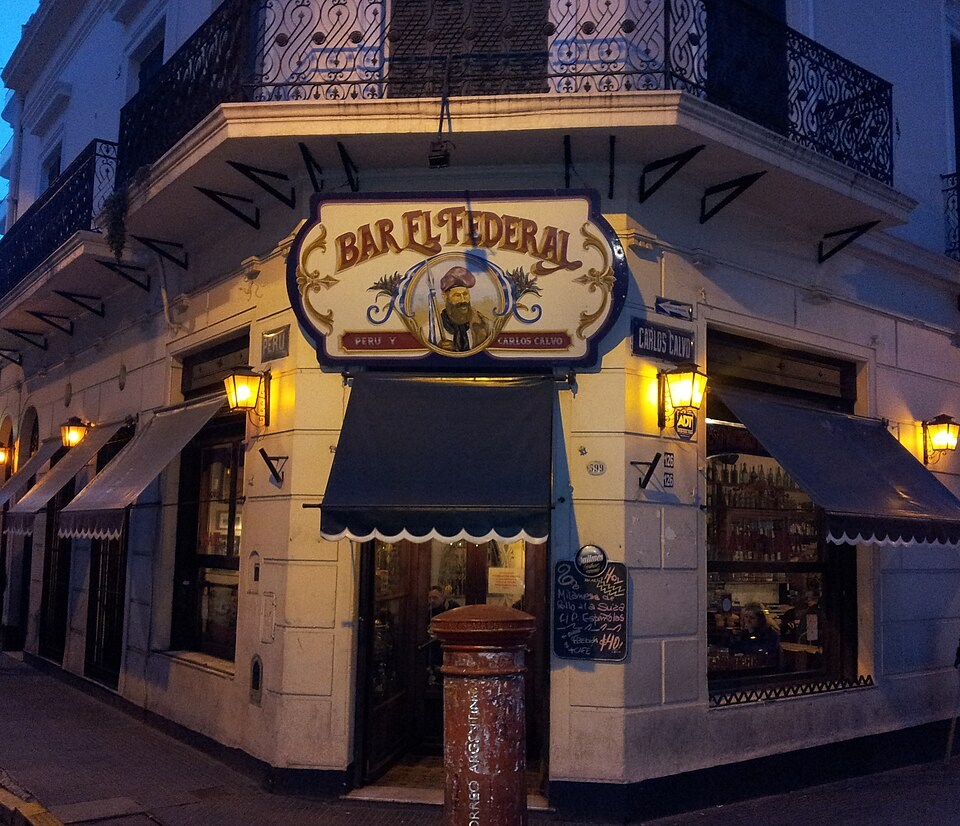 Fachada noturna do Bar El Federal, um prédio de esquina em Buenos Aires. O letreiro ornamentado exibe um homem barbudo com chapéu e armas. Toldos azuis escuros cobrem as janelas, e luminárias amarelas iluminam a rua. Um poste de correio vermelho antigo está na calçada