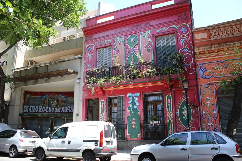 Fachada de um prédio rosa vibrante com detalhes ornamentais em verde, azul e rosa claro. Há um balcão com plantas e janelas com persianas. À esquerda, um prédio cinza com uma loja El Morocho del Abasto e carros estacionados na rua.
