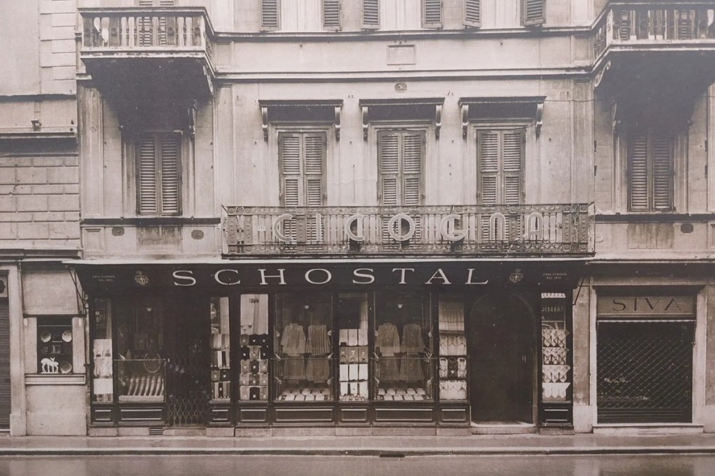 Fachada de um edifício antigo em sépia, com uma loja SCHOSTAL no térreo, exibindo roupas e acessórios em vitrines. Acima, um balcão com o nome CHIODINI e janelas com persianas fechadas. À direita, uma loja SIVA com porta de enrolar fechada