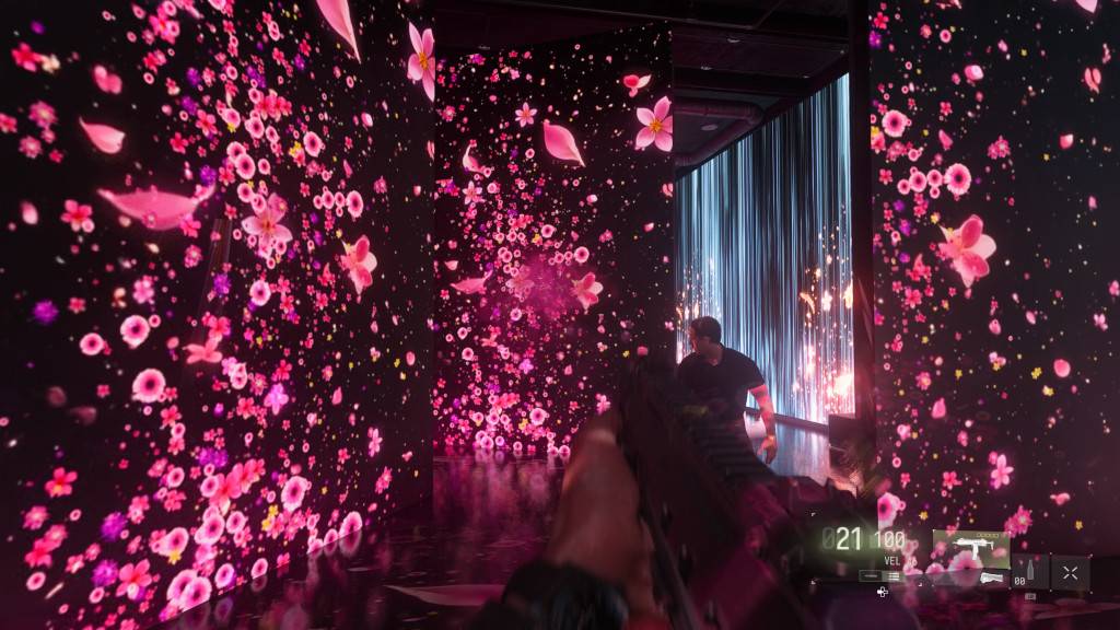 Cena de jogo com perspectiva em primeira pessoa segurando uma arma, em um ambiente escuro com paredes e chão refletindo projeções de flores e pétalas cor-de-rosa e roxas. Um homem de camiseta preta está ao fundo, à direita, em uma área iluminada por luzes verticais azuis e faíscas laranjas. Elementos da interface do jogo aparecem na parte inferior da tela.
