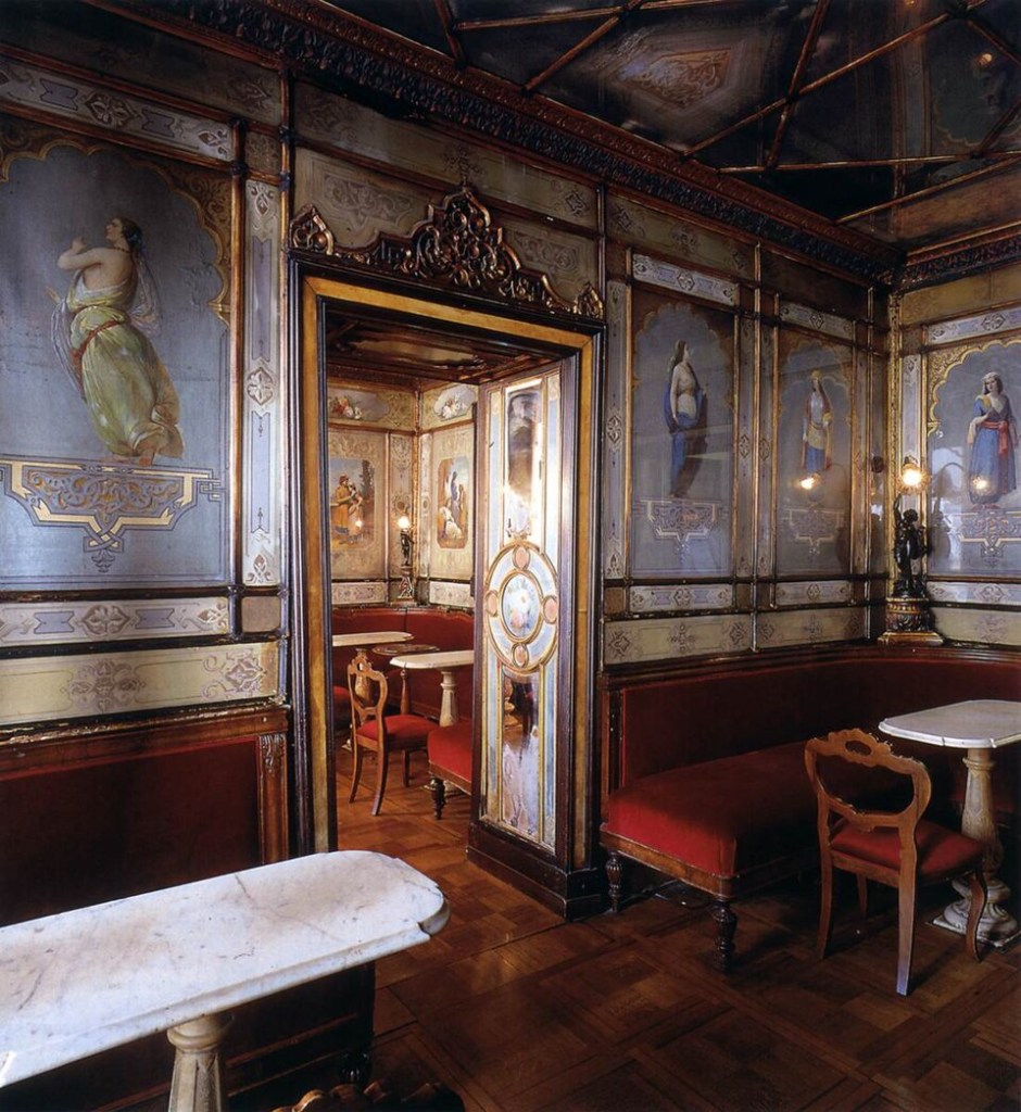 Salão luxuoso com paredes decoradas por pinturas de figuras femininas e detalhes dourados. Sofás vermelhos estofados e mesas de mármore branco, com cadeiras de madeira escura, preenchem o ambiente. O teto é ornamentado com relevos e o chão de madeira tem padrão geométrico. Uma porta entalhada leva a outro cômodo similar.