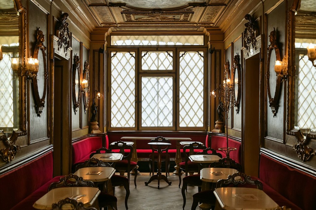 Interior de um café ou restaurante luxuoso, com paredes escuras e detalhes dourados. Há janelas grandes com grades em losango, sofás vermelhos de veludo e mesas pequenas com cadeiras ornamentadas. Espelhos e arandelas com luzes quentes decoram as paredes, criando uma atmosfera clássica e elegante