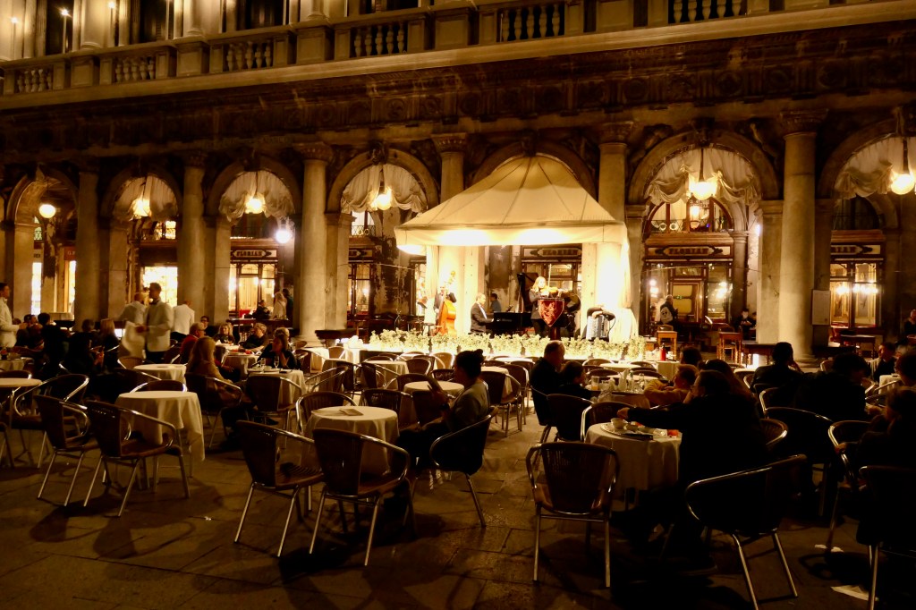 Pessoas sentadas em mesas de um café ao ar livre à noite, com um palco iluminado e músicos tocando ao fundo. O ambiente é elegante, com colunas e arcos de um edifício antigo, e garçons atendendo os clientes