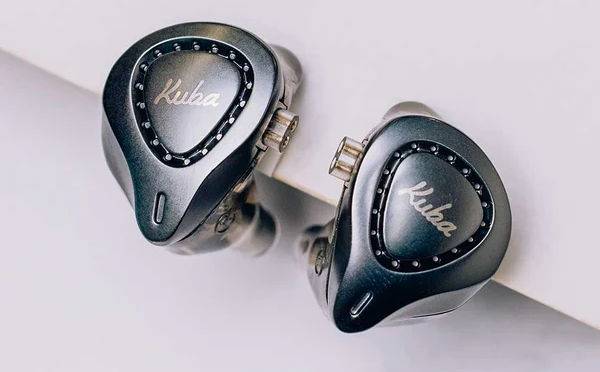 Dois fones de ouvido intra-auriculares cinza-escuros, com o nome Kuba gravado em branco no centro e um anel de pequenos pontos pretos ao redor, sobre uma superfície branca