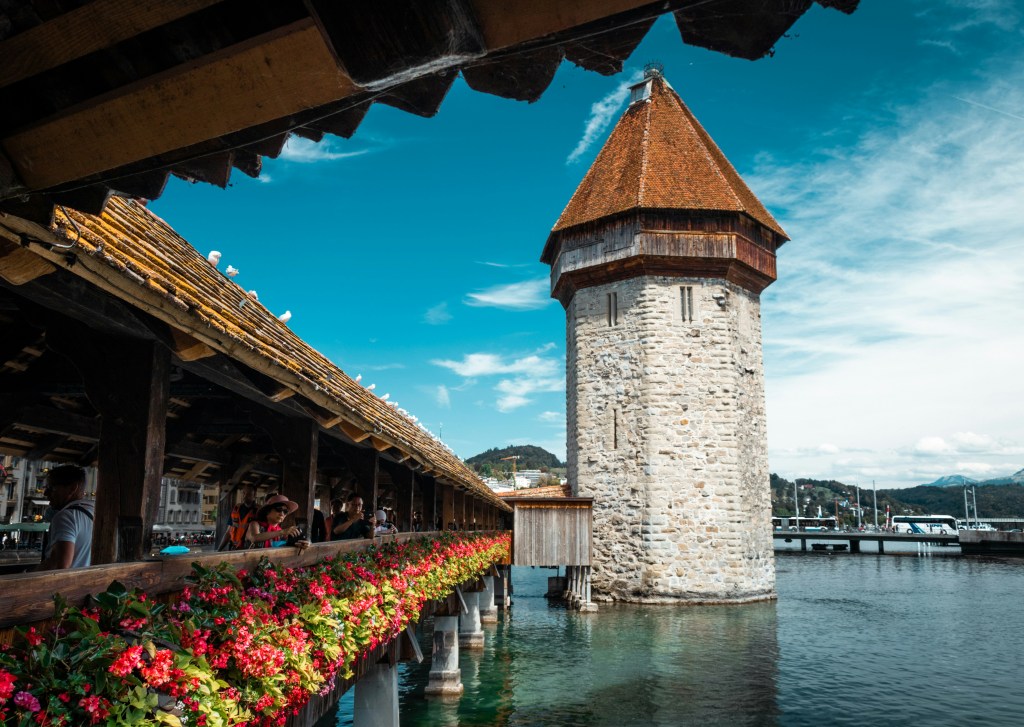 Ponte de madeira Kapellbrücke em Lucerna, Suíça, com telhado coberto por musgo e flores vermelhas. À direita, a torre de pedra Wasserturm com telhado marrom pontiagudo, sobre a água. Pessoas caminham pela ponte, e ao fundo, montanhas e um céu azul com nuvens brancas