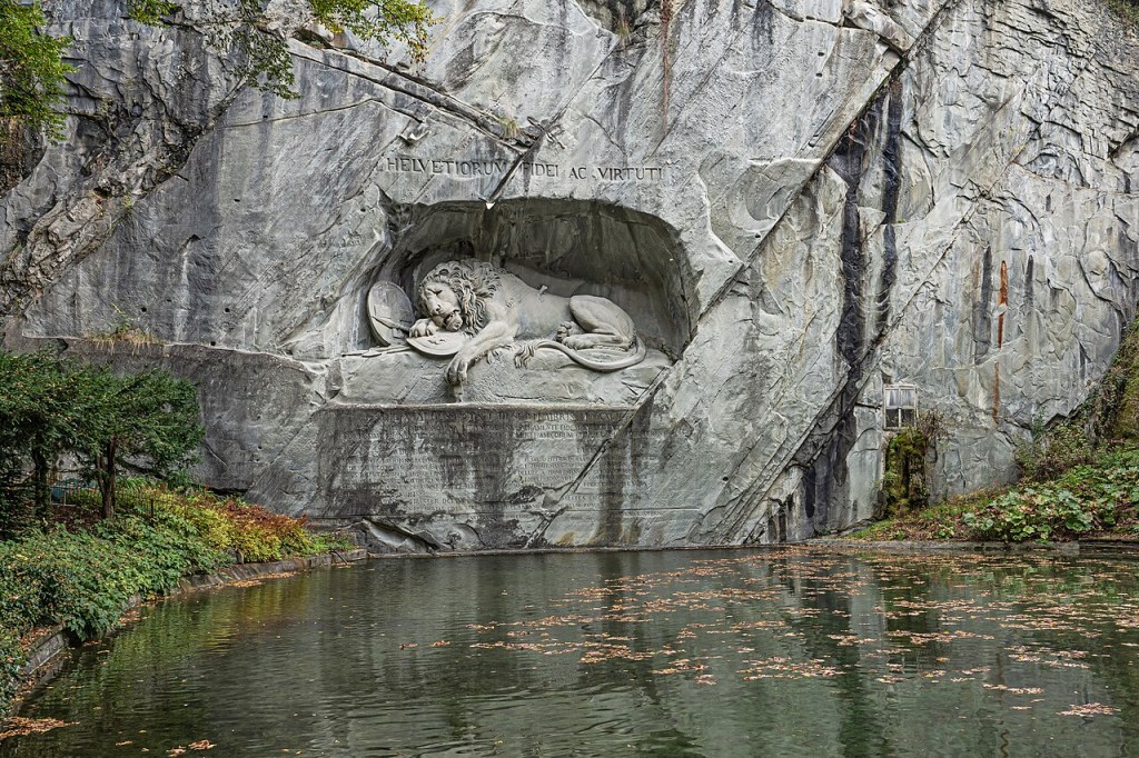 Escultura do Leão de Lucerna, um leão moribundo esculpido em rocha cinza, deitado sobre um escudo com a flor-de-lis, com uma lança quebrada em seu corpo. Acima, a inscrição HELVETIORUM FIDEI AC VIRTUTI. Abaixo, um lago escuro com folhas secas flutuando, e vegetação verde e marrom nas margens