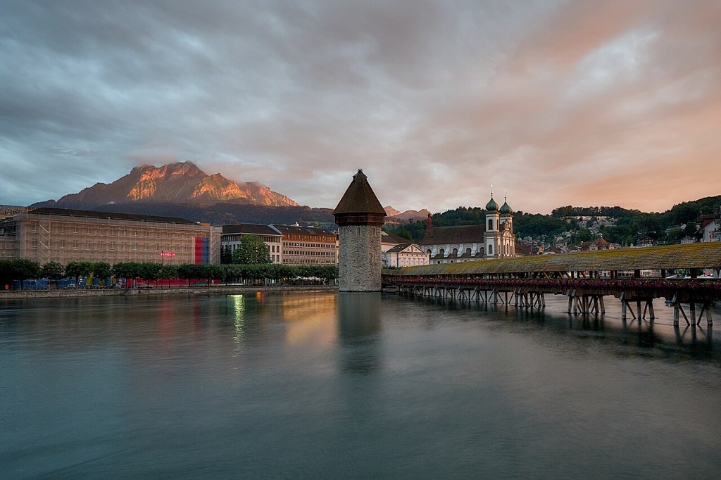 Vista panorâmica de Lucerna, Suíça, ao entardecer. O rio Reuss em primeiro plano reflete o céu nublado com tons alaranjados. À esquerda, edifícios e uma montanha com picos iluminados pelo sol poente. No centro, a torre de água medieval Wasserturm e a ponte de madeira Kapellbrücke, com a Igreja Jesuíta ao fundo, sob um céu dramático