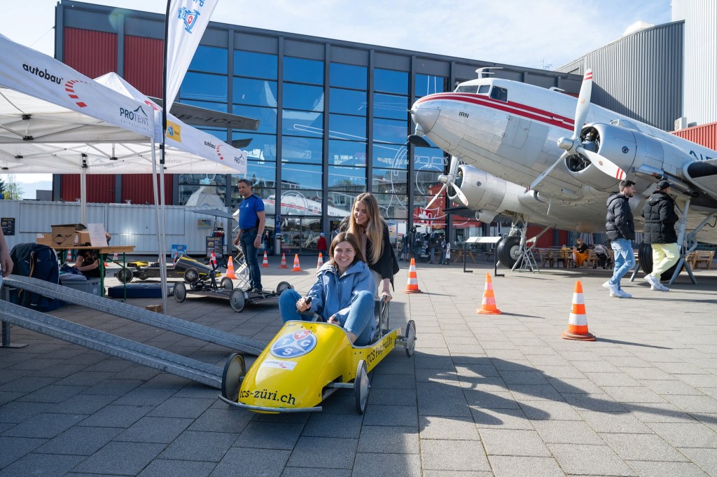 Duas mulheres sorridentes em um evento ao ar livre. Uma está sentada em um carrinho de rolimã amarelo, enquanto a outra a empurra. Ao fundo, um avião antigo prateado e um prédio moderno com janelas espelhadas.