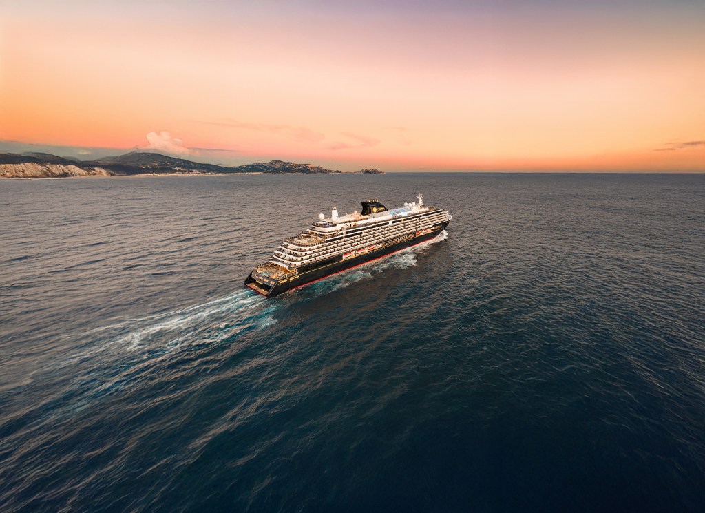 Navio de cruzeiro preto e branco navegando em águas azuis escuras, deixando um rastro de espuma. Ao fundo, montanhas costeiras e um céu em tons de laranja e roxo ao pôr do sol