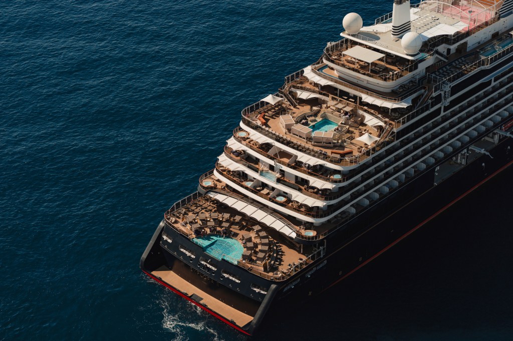 Vista aérea da popa de um navio de cruzeiro preto com detalhes vermelhos, navegando em águas azuis. Vários decks com piscinas, espreguiçadeiras e guarda-sóis brancos são visíveis
