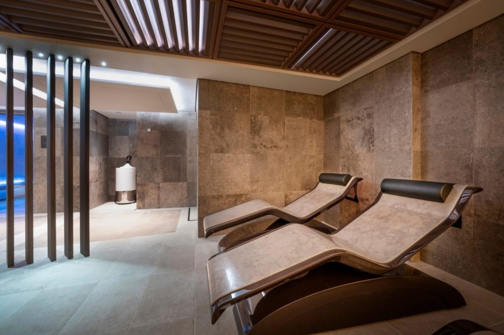 Ambiente de spa com duas espreguiçadeiras reclináveis bege e marrom, paredes de azulejo marrom texturizado e teto de madeira ripada. À esquerda, uma área de piscina com iluminação azul e divisórias verticais de madeira escura.