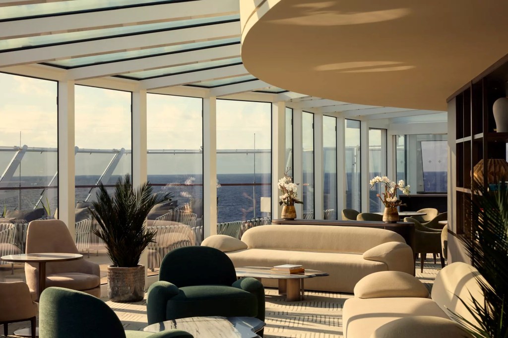 Lounge moderno e luxuoso em um navio, com janelas panorâmicas do chão ao teto revelando o oceano azul. Sofás e poltronas em tons de bege e verde-escuro, mesas de centro, plantas e arranjos florais, sob um teto de vidro e estrutura branca. A luz do sol entra pelas janelas, criando um ambiente acolhedor e sofisticado.
