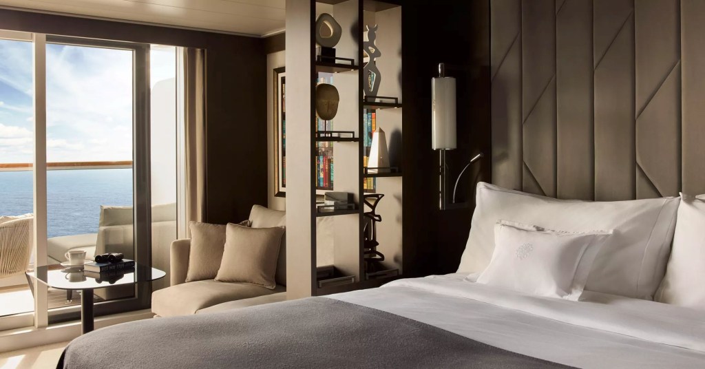 Quarto de hotel luxuoso com cama king-size, lençóis brancos e colcha cinza. À esquerda, uma porta de vidro abre para uma varanda com vista para o oceano azul. Uma mesa redonda com xícara de café e binóculos, e um sofá bege com almofadas. Uma estante com livros e objetos decorativos divide o ambiente.