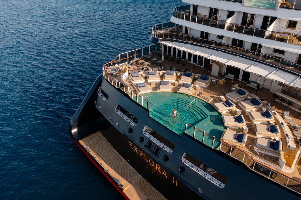 Vista aérea de um luxuoso navio de cruzeiro azul-marinho, EXPLORA II escrito em dourado. Uma mulher de biquíni branco está na piscina de borda infinita na popa, cercada por espreguiçadeiras e mesas. O mar azul profundo se estende ao redor.