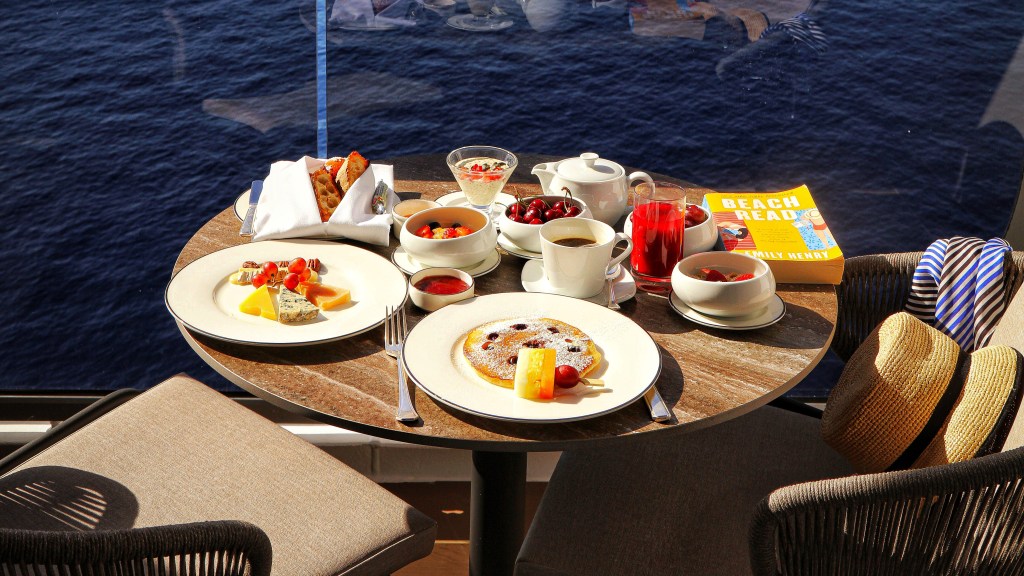 Mesa redonda com café da manhã farto em varanda de navio, com vista para o mar azul e céu claro. Há panquecas, frutas, pães, café, suco e um livro amarelo.