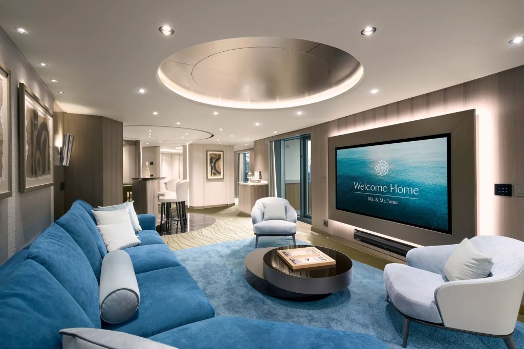 Sala de estar luxuosa com sofá azul-turquesa, poltronas claras, tapete azul e mesa de centro redonda. Uma grande TV exibe Welcome Home Ms. & Mr. Jones sobre um fundo de água. Iluminação embutida no teto e paredes, com um bar e balcão ao fundo