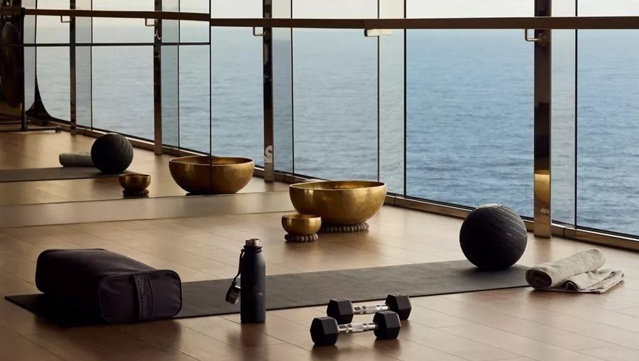 Estúdio de yoga com piso de madeira e janelas panorâmicas do chão ao teto, mostrando o oceano azul. Há tapetes de yoga, halteres, garrafa de água, almofada de meditação e tigelas tibetanas douradas.