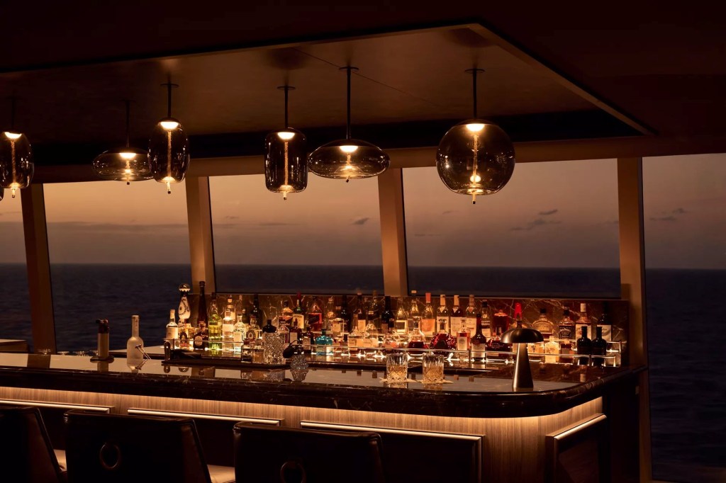 Bar de luxo em um navio, com balcão de mármore iluminado e diversas garrafas de bebidas. Quatro pendentes de vidro âmbar iluminam o ambiente. Ao fundo, janelas amplas revelam o oceano e um céu alaranjado no pôr do sol.