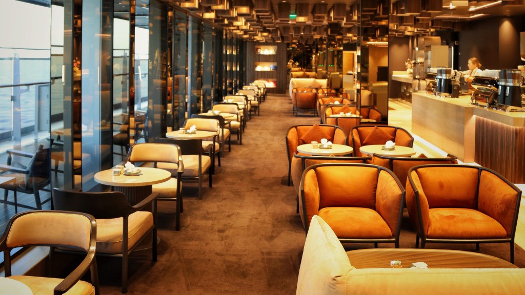 Interior de um café ou lounge moderno com iluminação quente. O teto possui painéis de madeira em relevo, criando um efeito tridimensional. À esquerda, janelas altas refletem o ambiente. Mesas redondas com cadeiras estofadas em tons de bege e laranja estão dispostas em fileiras sobre um carpete marrom. Ao fundo, um balcão de atendimento com uma pessoa.