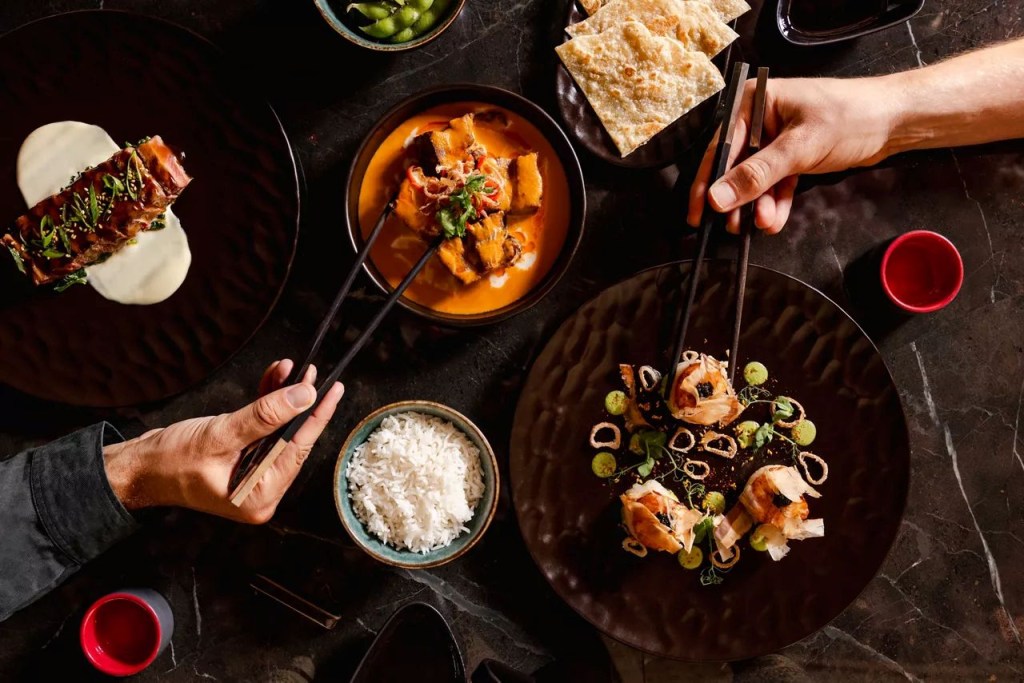 Duas mãos usando hashis para pegar comida em uma mesa escura com diversos pratos asiáticos: um filé com purê, um curry laranja, um bowl de arroz branco, edamame, pães achatados e um prato com rolinhos fritos e molho verde