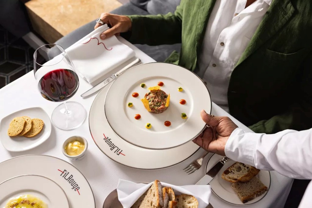 Garçom de camisa branca serve prato de tartare com molhos coloridos a cliente de blazer verde e camisa branca, sentado à mesa com taça de vinho tinto, pães e torradinhas