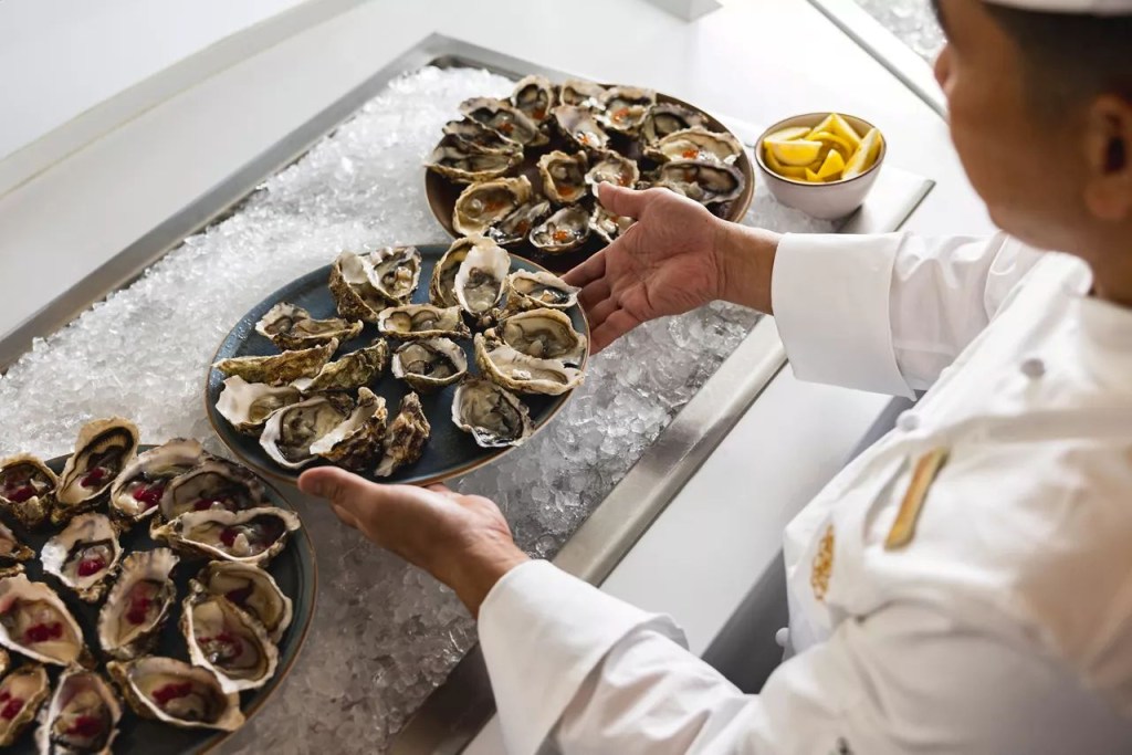 Chef de costas, vestindo dólmã branco, posiciona um prato oval com ostras frescas sobre uma cama de gelo picado. Outros dois pratos com ostras, algumas com ovas e outras com molho vermelho, estão visíveis. Uma tigela com fatias de limão completa a cena de um buffet de frutos do mar