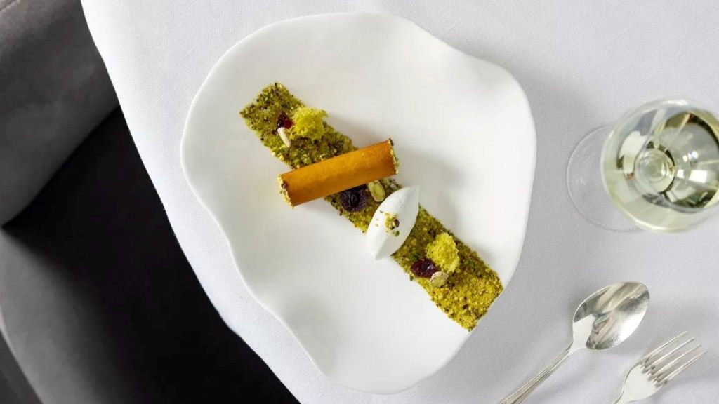 Sobremesa gourmet em prato branco ondulado, com base de pistache triturado, um rolinho dourado, quenelle de creme branco, frutas vermelhas e pistaches inteiros. Ao lado, taça de vinho branco, colher e garfo sobre toalha de mesa branca