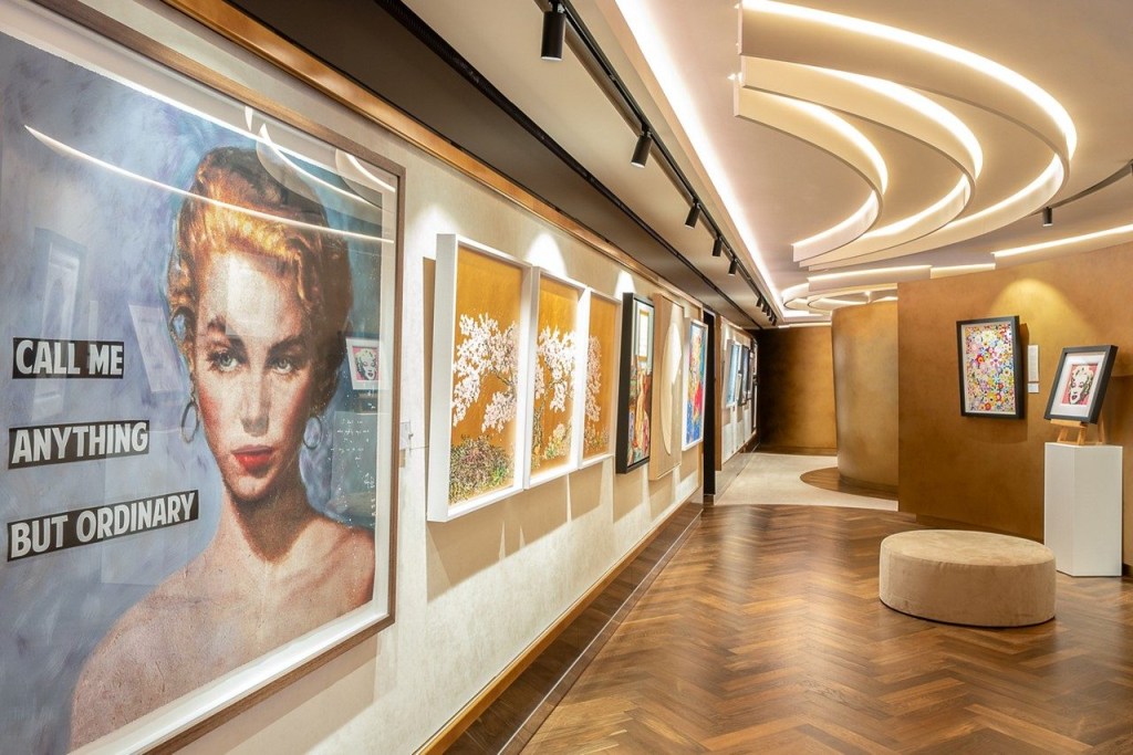 Corredor de galeria de arte com piso de madeira em espinha de peixe e teto iluminado. À esquerda, uma pintura de mulher loira com a frase CALL ME ANYTHING BUT ORDINARY. Outras obras de arte adornam as paredes, e um pufe bege está no centro.