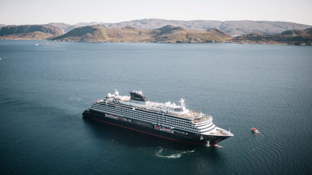 Navio de cruzeiro preto e branco, com vários andares e janelas, navegando em águas azuis escuras. Ao fundo, montanhas rochosas e um céu claro. Uma pequena embarcação laranja flutua à direita do navio.