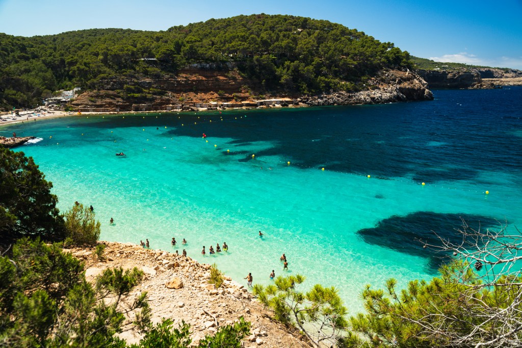 Vista aérea de uma praia com mar azul-turquesa e pessoas nadando, cercada por colinas verdes e rochosas sob céu claro. Boias amarelas delimitam a área de banho.