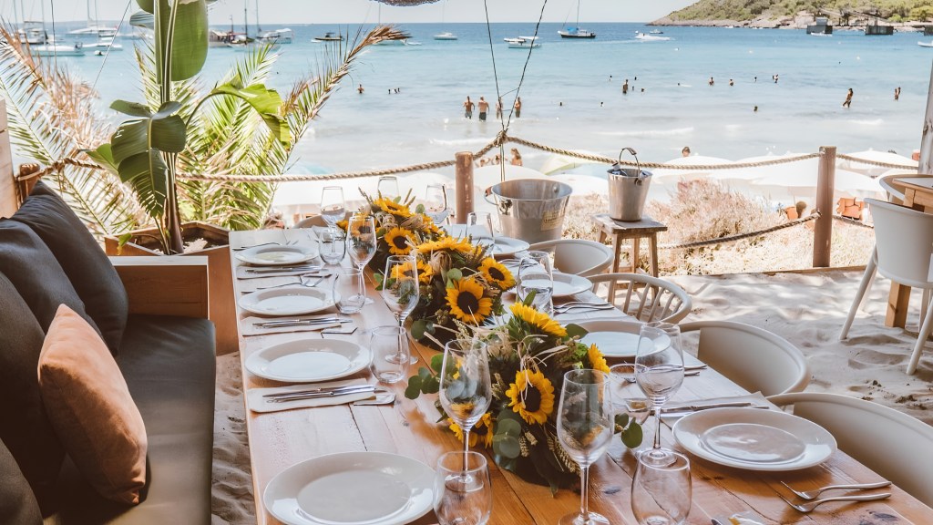 Mesa de madeira posta com pratos brancos, talheres e taças, decorada com arranjos de girassóis, em um restaurante à beira-mar com areia no chão. Ao fundo, pessoas nadam no mar azul-claro, com barcos ancorados e vegetação na costa