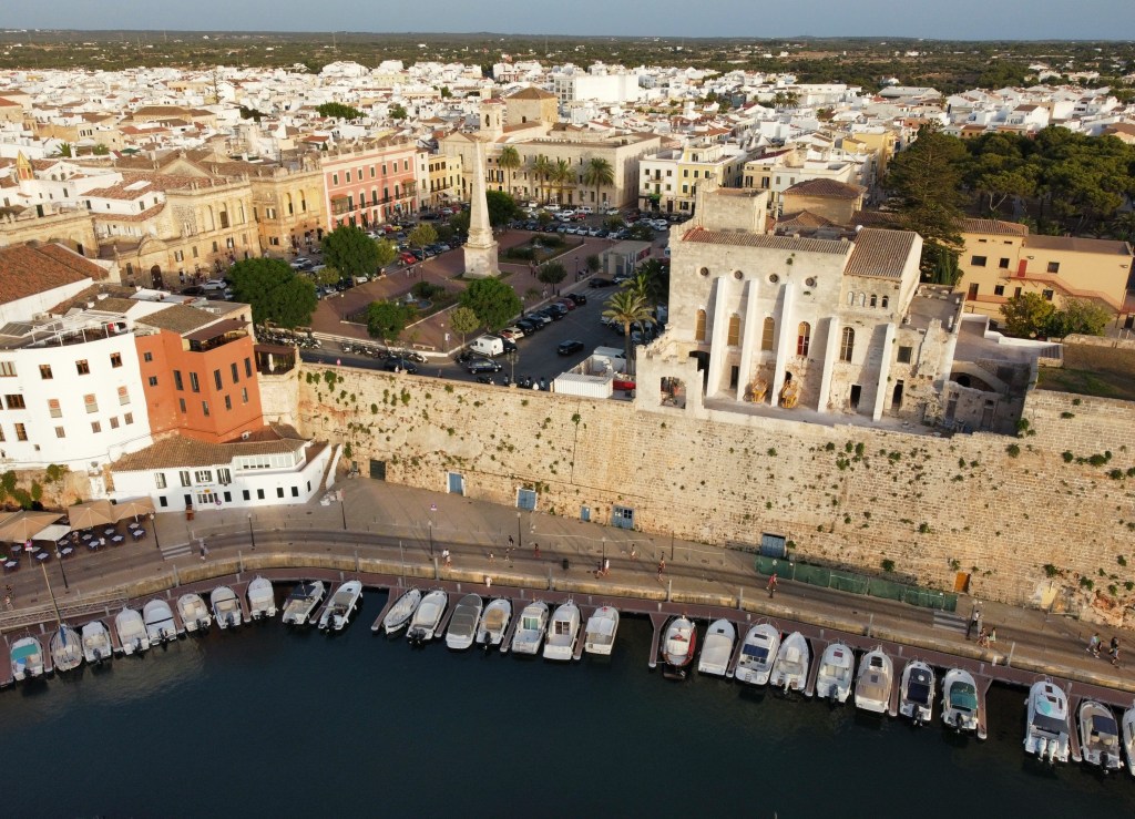 Vista aérea de uma cidade costeira com edifícios históricos de cores claras e telhados de terracota, um porto com barcos atracados e uma muralha de pedra antiga. No centro, uma praça com um obelisco e árvores.