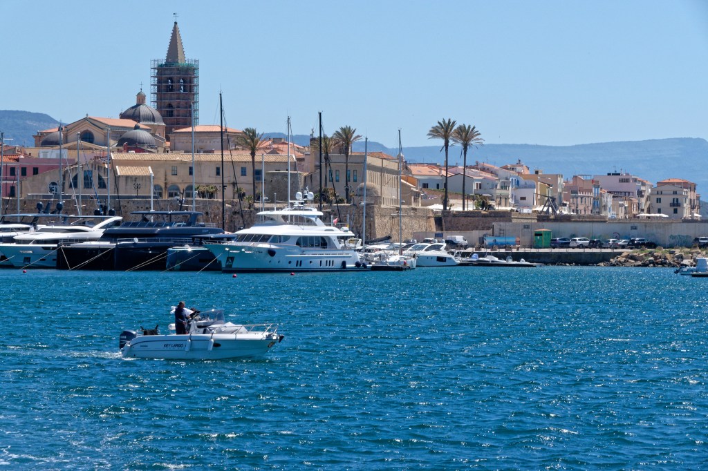 Homem em barco branco navegando em águas azuis, com porto de luxo e cidade costeira ao fundo, sob céu claro. Iates grandes e edifícios históricos com telhados de terracota e uma torre de igreja em destaque.