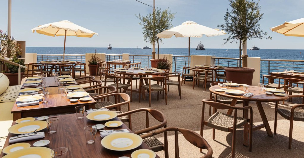Terraço de restaurante luxuoso à beira-mar, com mesas de madeira escura e cadeiras de palha. Pratos amarelos e talheres estão dispostos. Guarda-sóis bege oferecem sombra. O mar azul claro, com iates distantes, forma o cenário sob um céu ensolarado.