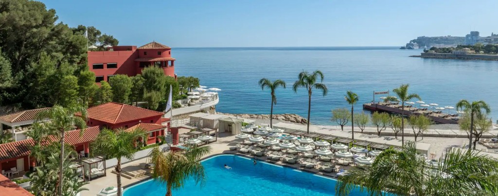 Vista aérea de um resort de luxo à beira-mar, com edifícios de telhado de terracota e paredes vermelhas, cercados por vegetação exuberante. Uma piscina azul brilhante com algumas pessoas nadando, espreguiçadeiras e guarda-sóis listrados na área da piscina e na praia de areia clara. Ao fundo, o mar azul calmo e um píer com mais espreguiçadeiras, sob um céu azul claro.